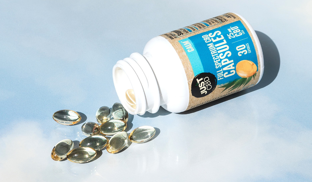 Open bottle of Just CBD full spectrum capsules with scattered capsules on a light blue background